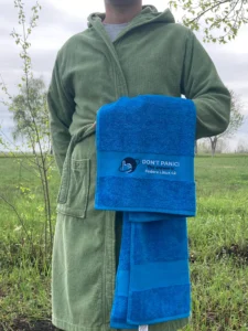 Gabor from Hello Tux, out standing in a field in a bathrobe holding his F42 towel. It says "Don't Panic — the answer is Fedora Linux 42"