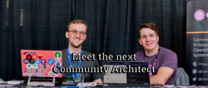 Two men sit behind laptops at a booth with large overlaid text reading, "Meet the next Community Architect". The background consists of black drapes and a partial event banner.