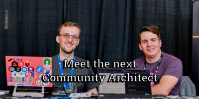 Two men sit behind laptops at a booth with large overlaid text reading, "Meet the next Community Architect". The background consists of black drapes and a partial event banner.